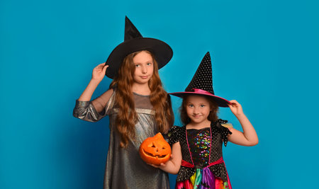 Two girls sisters in witches costumes with Halloween pumpkin holding hats and looking at camera on blue background.の写真素材