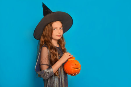 Cute Halloween child girl in witch costume with pumpkin looking at camera on a blue background with copy space. Wizard kid holding top of carved pumpkin or Jack o lantern pumpkin.の写真素材