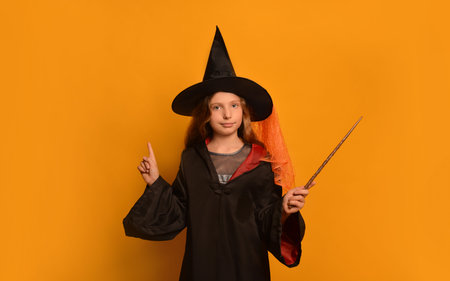 Cute pre-teen girl in wizard student costume holding magic wand or magic stick, looking at camera and holding index finger up on orange background.の写真素材