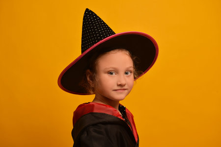 Portrait of little wizard girl in Halloween costume on yellow background. A kid wearing magic hat and mantle or robe and looking at camera. Happy Halloween and party concept.の写真素材