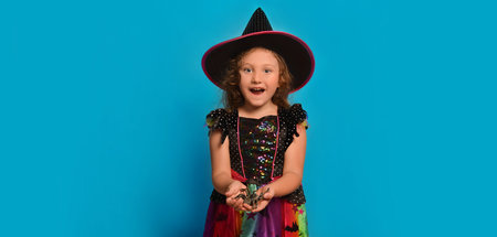 Surprised happy little girl wearing Halloween witch costume holding toy spider and looking at camera on a blue background with copy space for text. Kid in wizard dress costume.の写真素材