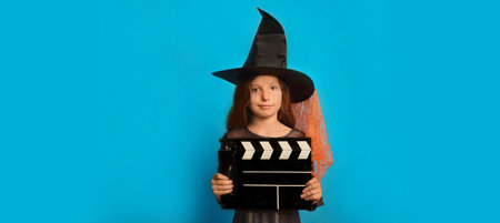 Little witch girl wearing wizard hat, holding classic black film making clapperboard and looking at camera on a blue background with copy space. Halloween horror cinema concept.の写真素材