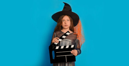 Scared little girl in witch costume holding film making clapperboard and looking at camera on blue background with copy space. I am afraid. Fright. Halloween movie concept.の写真素材