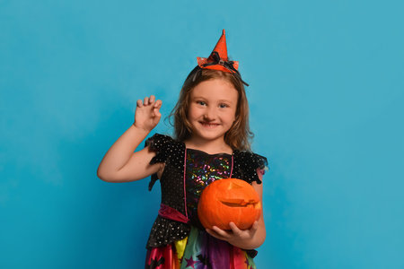 Little smiling witch girl showing Boo gesture and holding Halloween pumpkin or Jack o lantern pumpkin on blue background. Happy child in witch costume with hat looking at camera.の写真素材
