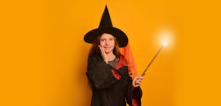 Happy smiling child girl in wizard student costume or black mantle robe holding hand on cheek with luminous magic wand or glowing magic stick on yellow background with copy space.の写真素材