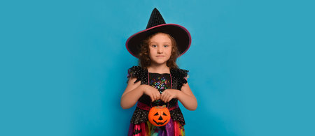 Cute little girl wearing Halloween witch costume or wizard costume, holding pumpkin candy basket or Halloween Jack o Lantern candy bucket and looking at camera on blue background.の写真素材