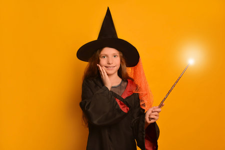 Happy smiling child girl in wizard student costume holding hand on cheek with luminous magic wand or glowing magic stick and looking at camera on yellow background.の写真素材