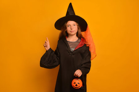 Little girl in wizard student costume holding finger up or pointing index finger up with Halloween Jack o Lantern pumpkin candy basket and looking at camera on a yellow background.の写真素材