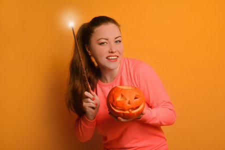 Happy young woman holding luminous magic wand or glowing magic stick with carved Halloween pumpkin or Jack o lantern pumpkin and looking at camera on yellow background.の写真素材