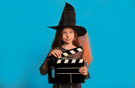 Little witch girl in Halloween wizard costume holding classic black film making clapperboard and looking at camera on a blue background. Cinema industry or horror movie concept.の写真素材