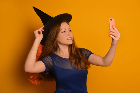 Young witch girl taking selfie photo on cellphone or smartphone and holding black hat on a yellow background. Wizard woman in blue dress doing selfie shot on mobile cell phone.の写真素材