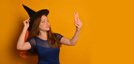Young witch girl taking selfie photo on cellphone or smartphone and holding black hat on yellow a background with copy space. Wizard woman doing selfie shot on mobile cell phone.の写真素材