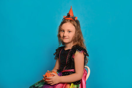 Cute little witch girl in Halloween dress costume sitting on a chair and holding Halloween pumpkin candy basket or Jack o Lantern candy bucket on a blue background.の写真素材