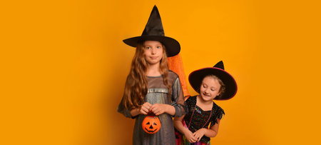 Two girls sisters dressed up as Halloween witches or wizards on yellow background with copy space. Kids in carnival costumes holding Jack o Lantern pumpkin candy basket.の写真素材