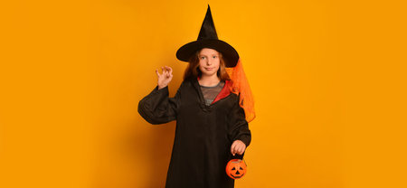 Witch girl in wizard student costume showing Boo gesture, holding Halloween pumpkin candy basket and looking at camera on yellow background with copy space. Trick or treat concept.の写真素材