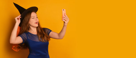 Young witch girl taking selfie photo on cellphone or smartphone and holding hat on yellow background. Halloween woman doing selfie shot on mobile phone and sticking tongue out.の写真素材