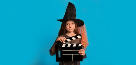 Little witch girl in Halloween costume holding classic black filmmaking clapperboard and looking at camera on blue background with copy space. Halloween horror movie concept.の写真素材