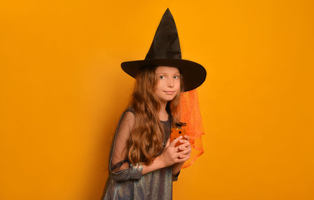 Witch kid girl holding polka dot festive glass with lemonade or juice, leaning forward and looking at camera on yellow background.の写真素材