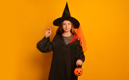 Little witch girl in wizard student costume showing Boo gesture, holding Halloween pumpkin candy basket or Jack o Lantern candy bucket and looking at camera on yellow background.の写真素材