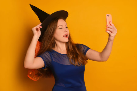 oung witch girl taking selfie photo on cellphone or smartphone and holding hat on yellow background. Halloween woman doing selfie shot on mobile phone and sticking tongue out.の写真素材