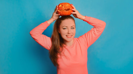 Happy young woman holding Halloween pumpkin or Jack o lantern pumpkin on head and looking at camera on blue background. Smiling girl in pink pullover celebrating Halloween.の写真素材