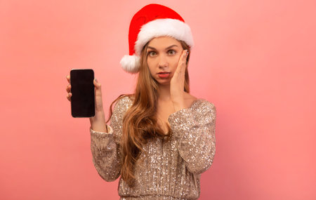 Surprised shocked girl in Santa Claus hat and sparkly dress holding hand on cheek, showing black screen of smartphone and looking at camera on pink background.の写真素材