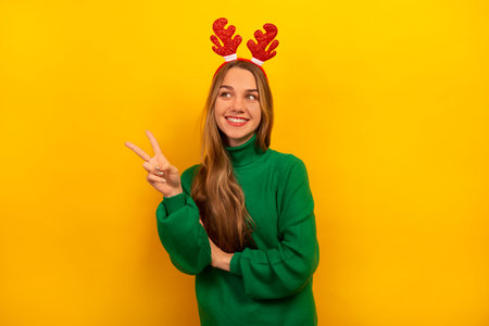 Happy smiling brunette girl showing peace v-sign gesture with two fingers, wearing green sweater with red Christmas toy reindeer antlers and looking up on yellow background.の写真素材