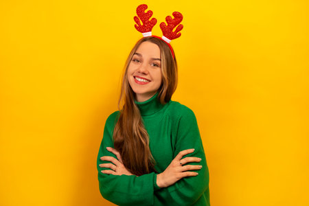 Happy laughing girl wearing green knitted sweater with red Christmas reindeer antlers and looking at camera on a yellow background. Young smiling woman standing with crossed arms.の写真素材