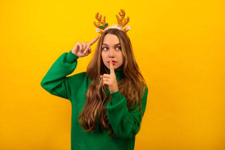 Young brunette woman showing silence gesture, pointing to golden Christmas reindeer antlers and looking up. Young woman wearing green sweater and standing on a yellow background.の写真素材