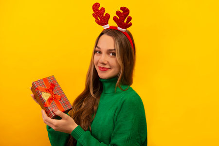 Happy smiling girl wearing red Christmas reindeer antlers and green knitted sweater, holding wrapped present or gift box and looking at camera on a yellow background.の写真素材