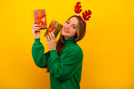 Happy cheerful young woman in Christmas reindeer antlers holding wrapped presents and looking at camera on yellow background. Smiling girl wearing green sweater and showing gifts.の写真素材