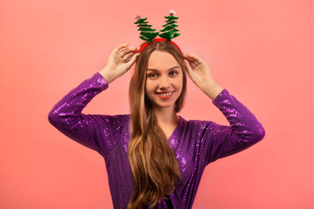 Happy smiling cheerful girl holding Christmas tree glasses headwear, wearing purple sparkly glitter dress and looking at camera on a pink background.の写真素材
