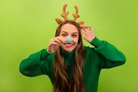 Happy smiling funny woman in Christmas reindeer antlers headwear and green sweater holding silver ball instead of nose on a colorful green background.の写真素材