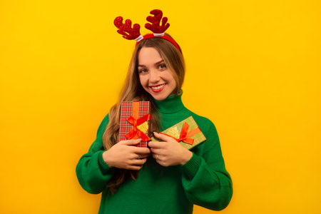 Happy smiling young woman holding wrapped presents or gift boxes, wearing red Christmas reindeer antlers with green knitted sweater and looking at camera on yellow background.の写真素材