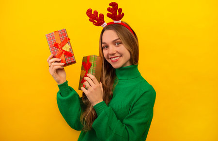 Happy smiling girl in red Christmas deer antlers showing wrapped presents and looking at camera on a yellow background. Girl wearing a green knitted sweater and holding gifts.の写真素材
