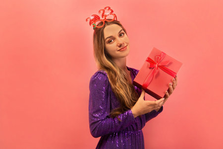 Cheerful young woman holding pink present or gift box with a bow, wearing purple sparkly dress with red party glasses and looking at camera on a pink background.の写真素材