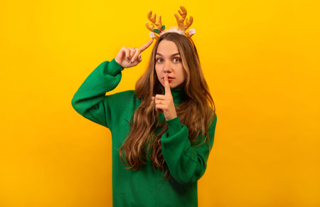 Brunette girl showing silence gesture, pointing to golden Christmas reindeer antlers and looking at camera. Young woman wearing knitted green sweater on a yellow background.の写真素材