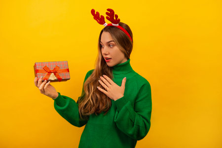 Surprised shocked girl holding wrapped present, keeping hand on chest and looking at gift on yellow background. Young woman wearing green sweater and Christmas reindeer antlers.の写真素材
