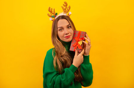 Beautiful brunette girl holding wrapped present or gift box, wearing green knitted sweater with Christmas toy reindeer antlers and looking at camera on a yellow background.の写真素材