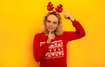 Happy young blonde woman showing silence gesture, pointing to red Christmas reindeer antlers and looking at camera on yellow background. Smiling girl wearing knitted red sweater.の写真素材