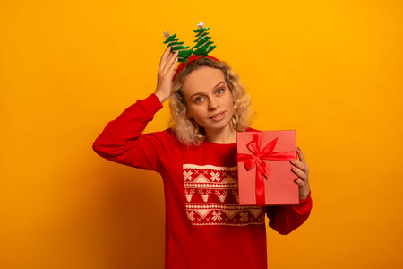 Young cheerful blonde woman wearing knitted red sweater with pattern ornament and party Christmas tree headwear looking at camera and holding pink present on a yellow background.の写真素材