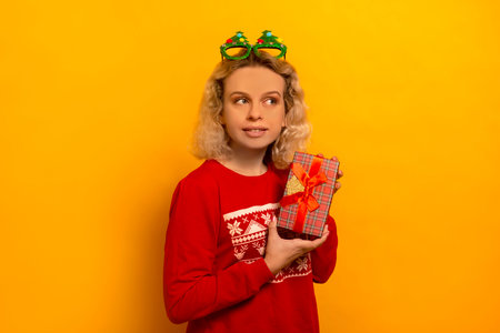 Young blonde woman holding Christmas gift or red wrapped present and looking aside on yellow background. Girl wearing red sweater and funny Christmas tree glasses.の写真素材
