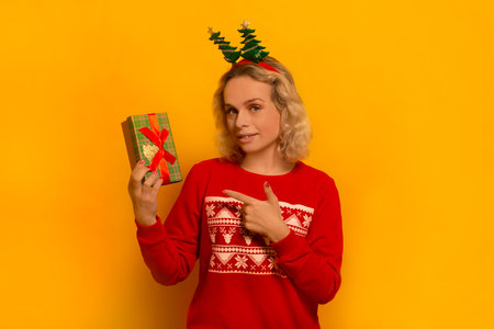 Young blonde woman in party Christmas tree headwear and knitted red sweater with pattern ornament pointing to wrapped gift box and looking at camera on a yellow background.の写真素材