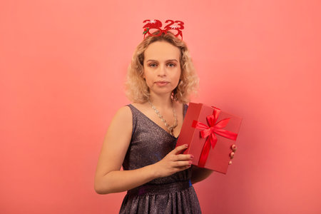 Disgruntled young woman holding present or gift, wearing silver sparkly glitter dress, necklace with party glasses 2023 and looking at camera on pink background.の写真素材