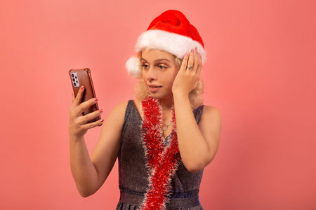 Surprised shocked young woman in Santa Claus hat, silver sparkly dress with red tinsel looking on smartphone screen on pink background. Girl taking selfie or reading text message.の写真素材