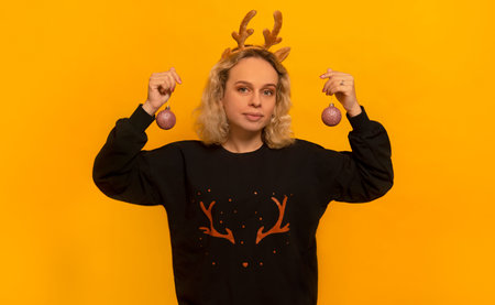 Young blonde woman in black sweater with pattern ornament and toy reindeer antlers holding Christmas balls and looking at camera on a yellow background.の写真素材