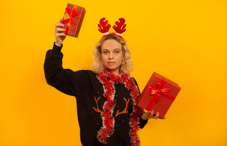 Young woman holding wrapped presents or gifts, wearing black sweater with deer antlers pattern, red Christmas reindeer antlers headwear and looking at camera on yellow background.の写真素材