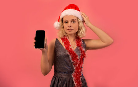 Pretty blonde girl in Santa Claus hat and silver sparkly dress with red Christmas tinsel showing black screen of smartphone and looking at camera on pink background. Mock up.の写真素材