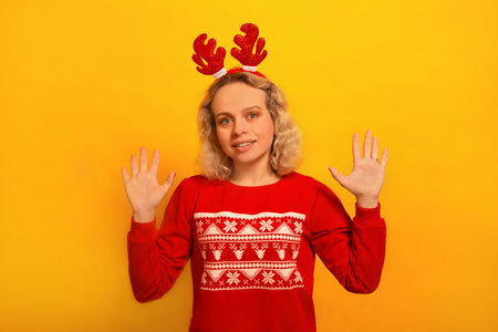 Happy smiling girl holding palms in front of her, wearing red knitted sweater with pattern and Christmas reindeer antlers headwear and looking at camera on yellow background.の写真素材
