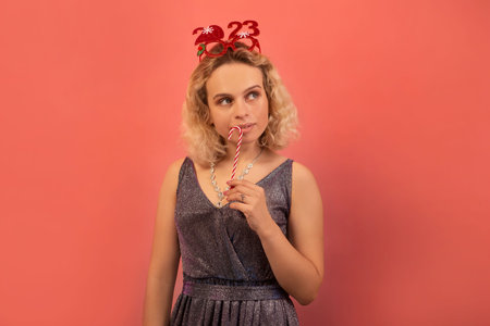 Young attractive blonde woman in silver sparkly glitter dress and red party glasses 2023 holding Christmas lollipop candy close to mouth and looking up on a yellow background.の写真素材